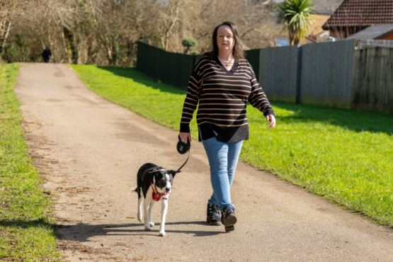 Woman walking a black-and-white dog on a paved trail beside a grassy verge on a sunny day, holding the leash in one hand and guiding the dog with the other.